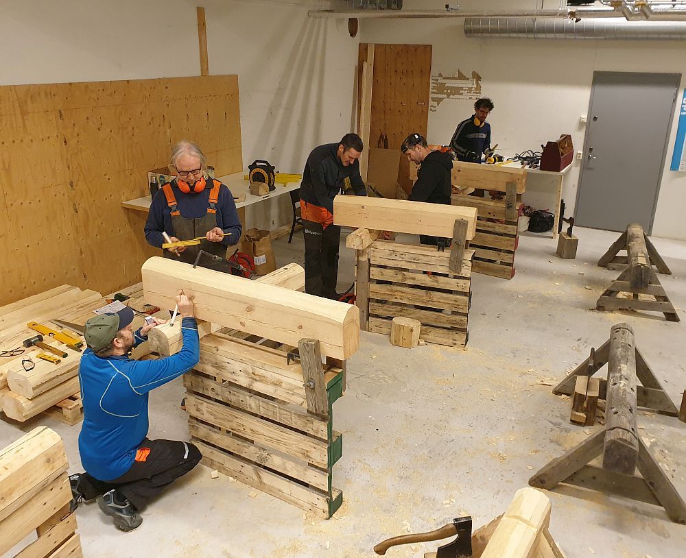 Kursdeltakere merker opp kantet tømmer på arbeidsbukker i et stort verksted.