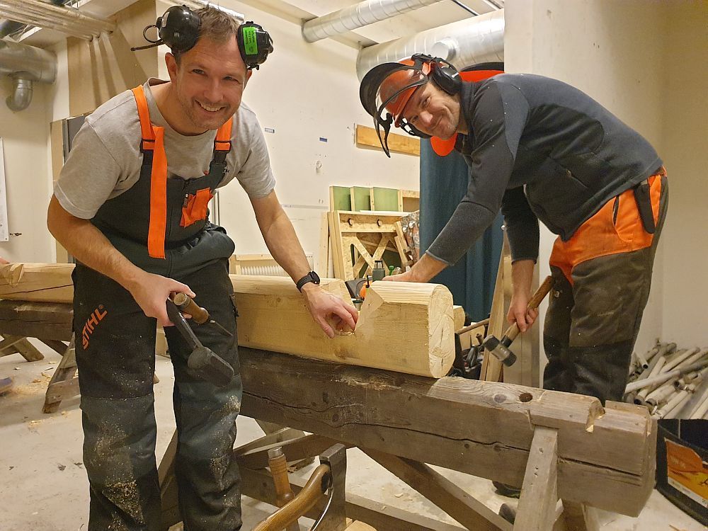 To smilende deltakere med hørselvern hogger en knute i en tømmerstokk.