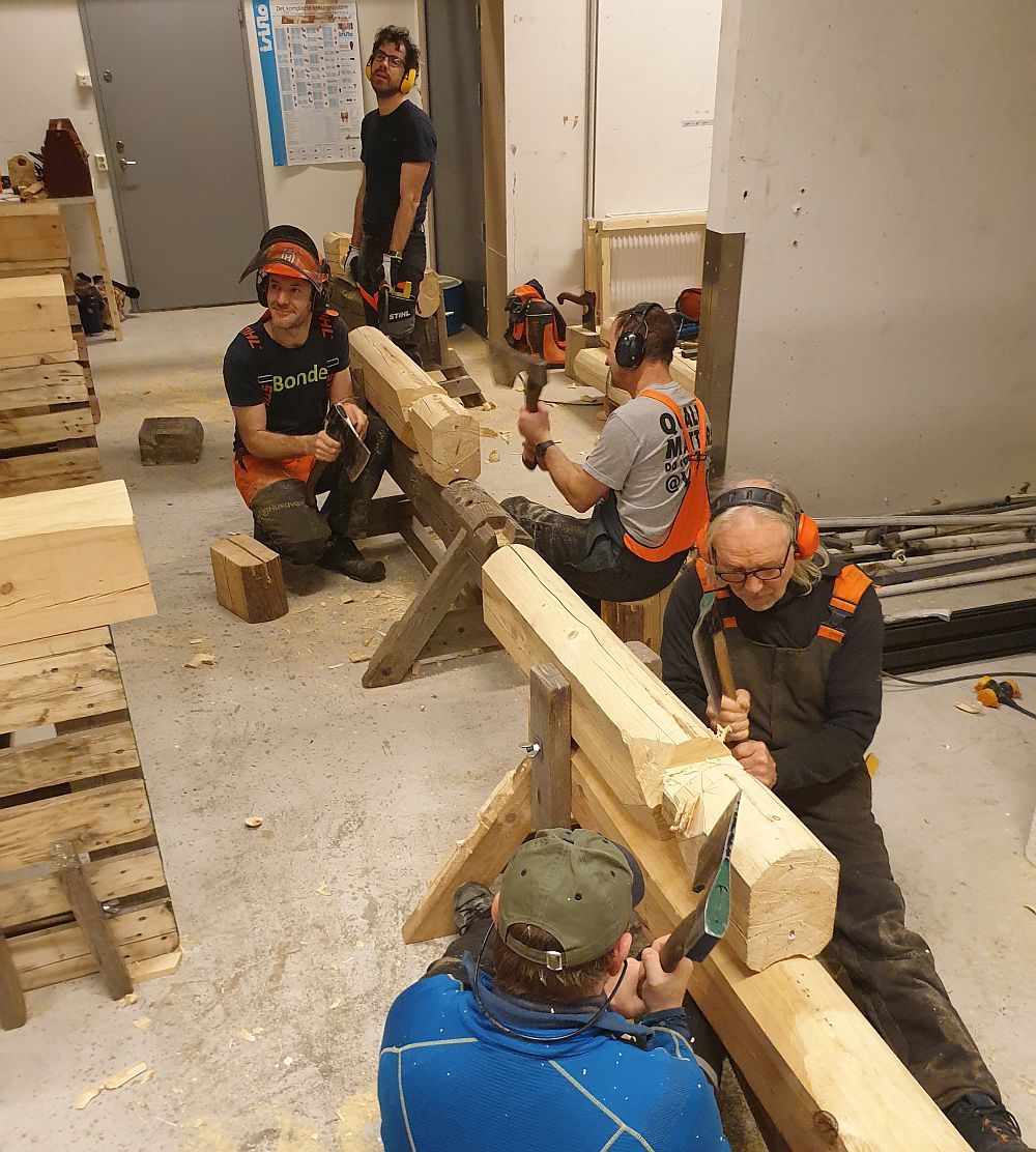 Fire deltakere hogger med øks på stokker som ligger på arbeidsbukker.